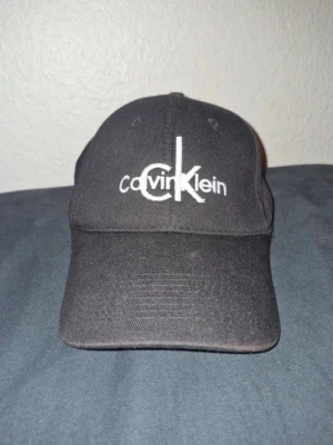 Svart keps från Calvin Klein - Säljer en svart keps från Calvin Klein med broderad logga framtill och 'ck' broderat baktill. Klassisk böjd skärm och justerbar rem med kardborre. Tillverkad i bomullstyg, perfekt för en clean och stilren look. 