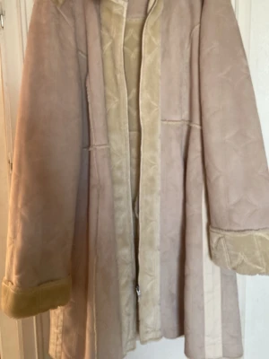 Beige fuskpälsfodrad kappa - Lång beige kappa med fuskpälsfoder och uppvikta ärmslut. Kappan har en mjuk yta i mockaimitation och detaljer i ljusare beige längs kanterna. Rak passform och knäppning framtill. Perfekt för kalla dagar och ger en stilren look.