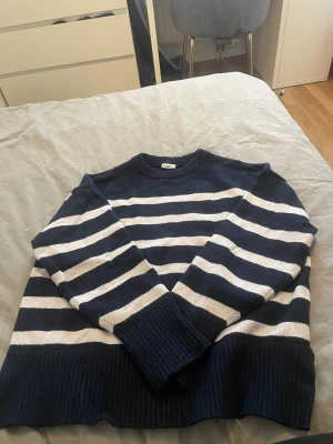 Marinblå randig stickad tröja Stories - Säljer en marinblå stickad tröja från Stories i storlek M. Tröjan har breda vita horisontella ränder, rund halsringning och ribbstickade muddar vid ärmslut och nederkant. Perfekt för dig som gillar klassisk och avslappnad stil.