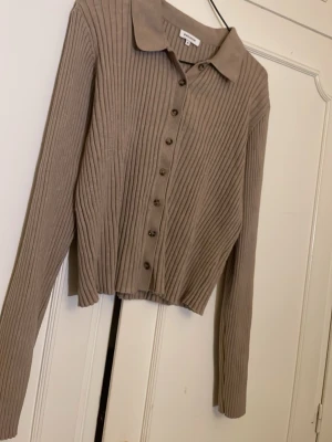 Beige ribbad kofta med krage - Snygg beige ribbad kofta med långa ärmar och klassisk krage. Koftan har knappar framtill och är något croppad i modellen. Perfekt att styla med jeans eller kjol för en trendig look.