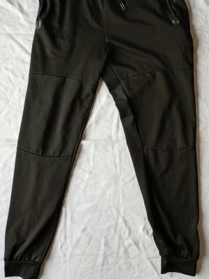 Svarta mjukisbyxor med dragkedjor - Säljer ett par svarta mjukisbyxor med dragkedjor vid fickorna och ribbade muddar vid bensluten. Byxorna har en relaxed fit och paneldetaljer över knäna för en sportig look. Perfekta för chill eller streetwear.