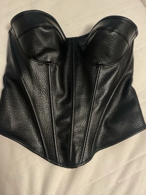 Svart korsett i fejkläder - Snygg svart korsett i fejkläder med markerade kupor och dekorativa sömmar framtill. Baksidan har snörning med metallöljetter för justerbar passform. Materialet är glansigt och ger en cool look. Perfekt för att lyfta vilken outfit som helst.