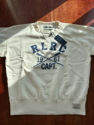 Vit sweatshirt från Polo Ralph Lauren - Aldrig använd Vit sweatshirt från Polo Ralph Lauren med blå tryckdetaljer framtill: RLRC, 1967 och CAPT. samt korsade åror. Rund halsringning och klassisk college-look. Perfekt för dig som gillar sportig och avslappnad stil. S, men sitter som S / M