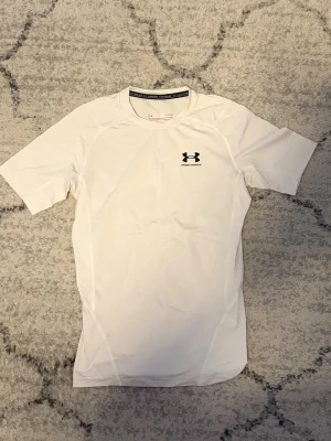 Under Armour  - Säljer en vit, tight och kortärmad träningströja från Under Armour. Tröjan har rund halsringning och svart logga på bröstet. Tillverkad i ett stretchigt funktionsmaterial som andas, perfekt för gym eller löpning.