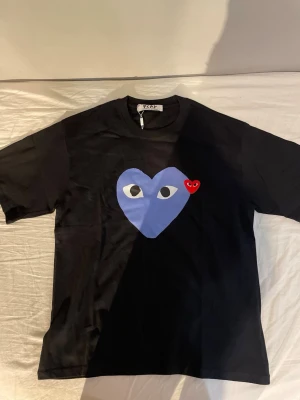Svart Comme des Garçons Play t-shirt - Svart t-shirt från Comme des Garçons Play med det ikoniska blå hjärtat med ögon och en liten röd hjärtdetalj på bröstet. Klassisk passform och rund halsringning. Tillverkad i mjuk bomull, perfekt för en avslappnad och trendig look.