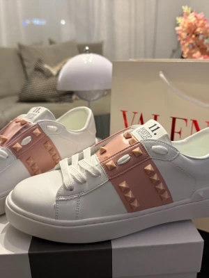 Valentino vita & rosa skor - Snygga vita sneakers från Valentino med rosa band och guldfärgade nitar över vristen. Skorna har klassisk låg profil, vita snören och är tillverkade i slätt läder. Perfekta för dig som vill sticka ut med en exklusiv touch.