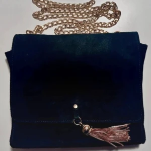 Axelremsväska i mocka med kedja - Två snygga axelremsväskor i läder , en i svart och en i beige. Båda har guldfärgad kedjerem och dekorativ tofs framtill. Perfekt för att lyfta din outfit med en trendig accessoar. Väskorna har enkel, fyrkantig form och stängs med lock.