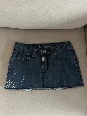 Mörkblå jeanskjol med detaljer - Kort mörkblå jeanskjol från J. Denim med rå kant nertill, dekorativa nitar / stenar på ena sidan och broderade detaljer på fickorna. Klassisk femficksmodell med knapp och dragkedja framtill. Perfekt för en trendig och cool  stil.