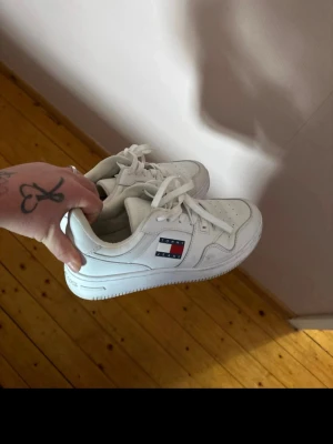 Vita Tommy Hilfiger sneakers - Snygga vita sneakers från Tommy Hilfiger med klassisk logga på sidan. Skorna har lågt skaft, perforerad tå och vita snören. Tillverkade i läderliknande material med detaljer i rött och blått på loggan. Perfekta för en clean och stilren look. Finns lite ytligt smuts men har inte provat att få bort de 