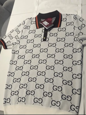 Polo Tshirt GG heöt ny - Säljer en vit polotopp från Gucci med svart GG-logga över hela plagget. Kragen är svart med röd orange detaljer, och toppen har korta ärmar med matchande kantband. Perfekt för dig som gillar exklusiv och trendig stil. storlek xl helt ny med ettiket