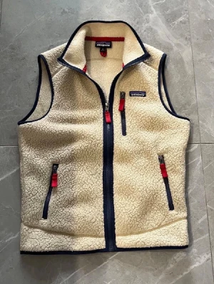 Beige fleeceväst från Patagonia - Snygg beige fleeceväst från Patagonia med marinblå detaljer och röda dragkedjor. Västen har hög krage, tre fickor med dragkedja och är gjord i mjukt teddyfleece-material. Perfekt för lager-på-lager och chill stil.