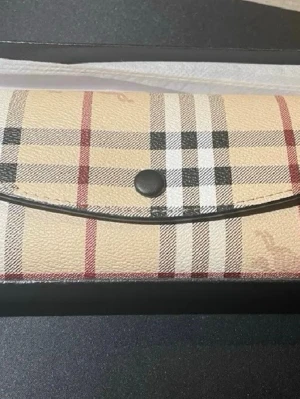 Beige plånbok med rutigt mönster - Snygg beige plånbok från Burberry med det klassiska rutiga mönstret i svart, vitt och rött. Plånboken har en avlång form och stängs med en knapp framtill. Tillverkad i tåligt konstläder med lyxig känsla och levereras i en svart ask.