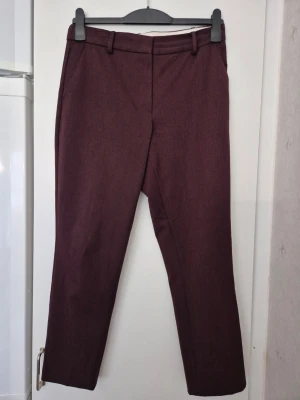 Kostymbyxor - Snygga vinröda/bruna kostymbyxor med raka ben och klassisk passform. Byxorna har bälteshällor, sidofickor fram och en passpoalficka bak. Perfekta för en stilren och modern look. Materialet ser ut att vara en mjuk och lite stretchig blandning. Gul fläck.