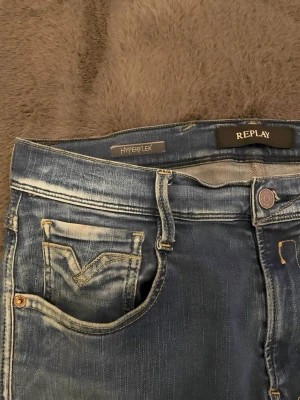 Blå Replay Hyperflex jeans - Säljer ett par blå slim fit jeans från Replay, modell Hyperflex. Jeansen har slitna detaljer, fem fickor och stretchigt material för extra komfort. Snygg tvätt med ljusare partier på låren och klassisk knappgylf.