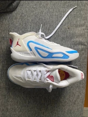 Jordan vita och blå låga sneakers - Säljer ett par fräscha sneakers från Jordan i vitt meshmaterial med blå detaljer och röd logga på sidan. Skorna har en sportig siluett, vita snören och en stabil sula. Perfekta för dig som gillar streetwear och basketinspirerad stil.