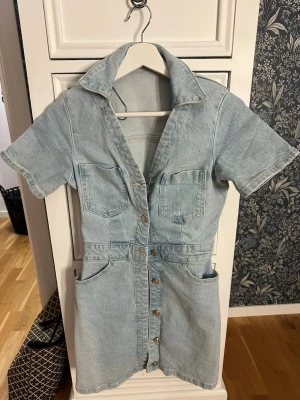 Kort jeansklänning med knappar - Snygg ljusblå jeansklänning med korta ärmar, stora fickor fram och bak samt knappar hela vägen framtill. Klänningen är helt oanvänd med prislappen kvar!☺️