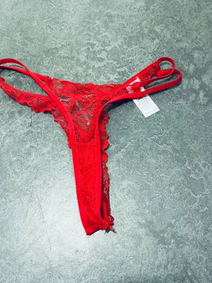 Röda spets-trosor med strassdetalj - Säljer ett par röda stringtrosor i spets med dekorativa strassdetaljer framtill. Trosorna har dubbla tunna band i midjan och ett feminint blommigt spetsmönster. Perfekt för dig som gillar detaljer och en sexig look.