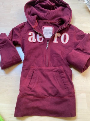 Vinröd hoodie från Aeropostale - Snygg vinröd hoodie från Aeropostale med halv dragkedja och stor magficka. Broderad text 'aero' framtill i vitt och rosa. Tillverkad i mjuk bomullsblandning med huva och ribbade muddar. Perfekt för en chill och avslappnad stil. Kan finnas lite missfärgning från solen men inget super super tydlgig 