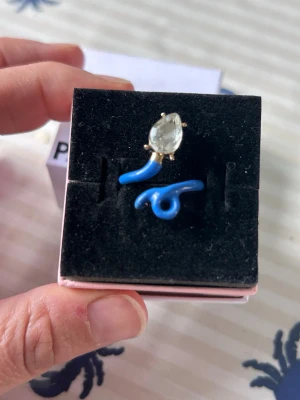 Blå ring med klar sten - Ring  med en böjd, blå metallform och en stor, droppformad klar sten i toppen. Ringen har en modern och lekfull design som verkligen sticker ut. Perfekt för dig som gillar färg och vill ha något annorlunda i din smyckessamling.