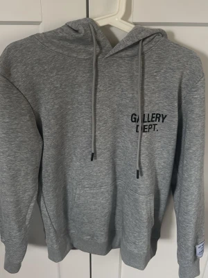 Grå hoodie från Gallery Dept. - Snygg grå hoodie från Gallery Dept. med svart tryck på bröstet och stor logga på ryggen. Klassisk modell med huva och dragsnören samt magficka framtill. Perfekt för en avslappnad streetwear-look.
