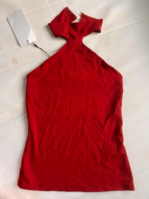 Röd one shoulder topp med knappar - Snygg vinröd topp med hög hals. Toppen är ärmlös och har en tight passform som framhäver siluetten. Tillverkad i mjukt material som känns skönt mot huden. Färgen liknar mest bilden 2.