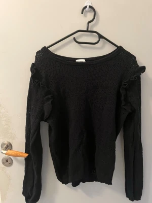 Svart stickad tröja med volang - Snygg svart stickad tröja med volangdetaljer längs axlarna och ärmarna. Tröjan har ett diskret hålmönster och rund halsringning. Perfekt för dig som gillar en enkel men ändå trendig look.