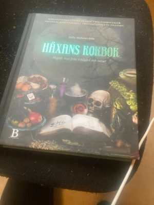 Häxans kokbok av Hella Nathorst-Böös - En magisk kokbok med recept och inspiration från trädgård och natur, skriven av Hella Nathorst-Böös. Omslaget är mörkt med mystiska illustrationer av växter, frukter och en dödskalle. Perfekt för dig som gillar natur, magi och matlagning.