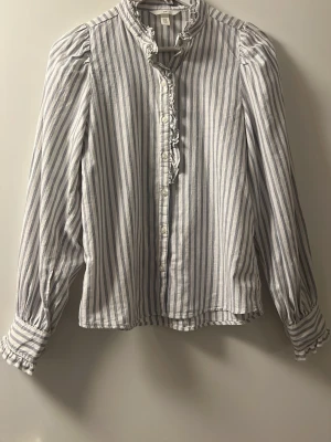 Randig blus med volangdetaljer - Snygg blus från H&M med vertikala blå och vita ränder. Blusen har volangdetaljer vid kragen och knäppningen, samt puffiga långärmar med knappar vid manschetterna. Tillverkad i ett lätt bomullstyg som ger en luftig känsla.