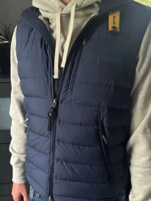 Mörkblå dunväst från Parajumpers - Snygg mörkblå dunväst från Parajumpers med quiltad design, hög krage och dragkedja framtill. Västen har två fickor med dragkedja och en gul detalj vid halsen. Perfekt för lager på lager och streetwear-stil. Kan bytas mot annan väst exempel Patagonia, hör av dig vid minsta fråga eller fundering!