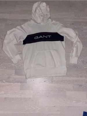 Vit hoodie från GANT med logga - Snygg vit hoodie från GANT med stor svart panel och röd kant över bröstet där GANT-loggan är tryckt. Klassisk känguruficka framtill och huva. Tillverkad i mjukt material, perfekt för en chill och avslappnad stil.