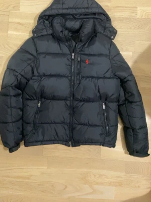 Svart dunjacka från Polo Ralph Lauren - Snygg svart dunjacka från Polo Ralph Lauren med huva och dragkedja framtill. Jackan har två sidofickor med dragkedja och en röd logga broderad på bröstet. Puffig modell som håller dig varm under kalla dagar. Perfekt för vintern!