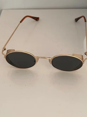 Runda solglasögon med guldfärgade bågar - Snygga solglasögon med tunna guldfärgade metallbågar och runda, mörka glas. Skalmarna har sköldpaddsmönstrade detaljer i brunt och orange. Perfekt accessoar för en trendig look.