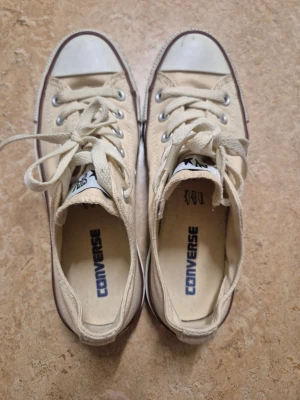 Beige Converse låga sneakers 37 - Klassiska låga Converse sneakers i beige canvas med vita gummisulor och tåhätta. Skorna har vita snören, silverfärgade öljetter och den ikoniska Converse-loggan på plösen och innersulan. Passar perfekt till jeans eller shorts för en avslappnad stil.