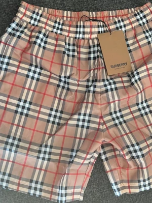 Rutig Burberry bad shorts - Säljer ett par klassiska Burberry shorts i beige med det ikoniska rutiga mönstret i svart, vitt och rött. Shortsen har elastisk midja och är perfekta för en trendig och avslappnad stil. Passar dig som vill ha något stilrent och tidlöst.