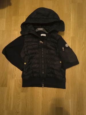 Svart dunjacka med huva från Moncler - Snygg svart dunjacka från Moncler med stor huva och dragkedja framtill. Jackan har ribbade ärmar, två fickor med tryckknappar och Moncler-logga på ena ärmen. Perfekt för kalla vinterdagar och riktigt stilren design.