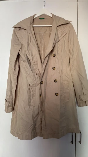 Beige trenchcoat med stora knappar - Stilren beige trenchcoat med dubbelknäppning och stora marmorerade knappar. Jackan har bred krage och två fickor framtill. Tillverkad i ett lätt tyg som passar perfekt för lager på lager. Klassisk och tidlös design. Jackan saknar dock skärp så säljs billigt