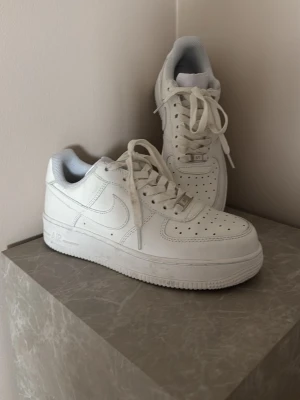 Nike Air Force 1 vita sneakers - Klassiska helvita Nike Air Force 1 sneakers i lågt utförande. Skorna har en stilren design med perforerad tå, vita skosnören och Nike swoosh på sidan. Tillverkade i slätt läder med en robust gummisula och diskreta detaljer i silver på snörningen. 38,5