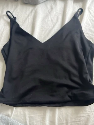 Svart linnetopp från Sisu - Stilren svart linnetopp från Sisu med smala axelband och v-ringad halsringning. Toppen har en croppad passform och är tillverkad i ett mjukt, lätt material som känns skönt mot huden. Perfekt att matcha med jeans eller kjol.
