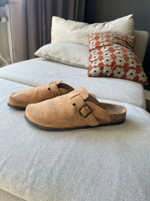 Birkenstock Boston - Säljer ett par klassiska bruna tofflor som liknar Birkenstock boston i mjuk mocka med justerbart spänne och korksula. Yttersulan är i slitstark gummi med bra grepp. Perfekta för chill hemma eller snabba ärenden utomhus. Ai gjorde beskrivning 