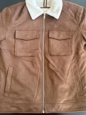 Brun jacka med fuskpäls-krage - Snygg brun jacka från LCW Vision i mockaimitation med en stilren vit fuskpäls-krage som ger en tydlig retroinspirerad look. Perfekt för dig som vill sticka ut med något unikt och trendigt.  Detaljer: 	•	Mockaimitation med mjuk känsla 	•	Vit fuskpäls-krage 	•	Två stora bröstfickor med lock 	•	Dragkedja framtill 	•	Sidofickor för extra funktion  Storlek: Passar M–L  En perfekt jacka för både vardag och stilrena outfits med retro vibe.  💬 Hör av dig vid intresse!  