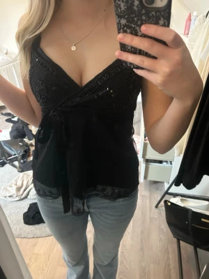 Svart omlottopp med pärlor och knyt - Snygg svart omlottopp med tunna axelband, dekorerad med pärlor och broderade detaljer. Toppen har knyt i midjan och mesh-tyg med fina detaljer längst ner.