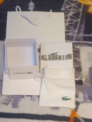 Silverfärgat armband från Lacoste - Snyggt armband från Lacoste i silverfärgad metall med länkdesign och diskreta mönstrade detaljer på länkarna. Kommer med original ask och tygpåse med Lacoste-logga. Perfekt accessoar för dig som gillar stilrena smycken med en sportig touch.