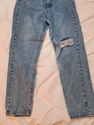 Blå mom jeans med slitning från Gina Tricot - Säljer ett par ljusblå mom jeans med hög midja och avslappnad passform. Jeansen har en tydlig slitning med hål på ena låret och klassisk femficksdesign. Tillverkade i denim med vintagekänsla och raka ben. Perfekta för en avslappnad och trendig look.
