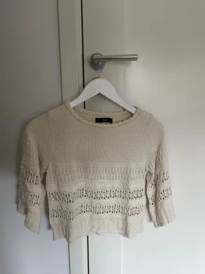 Beige stickad tröja med hålmönster - Säljer en beige stickad tröja med rund halsringning och trekvartsärmar. Tröjan har snygga hålmönster i ränder över hela plagget och en avslappnad passform. Perfekt för lager på lager och en chill look.