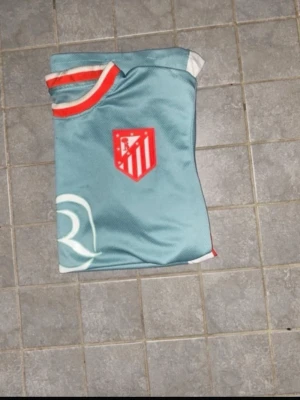 Atlético Madrid fotbollströja turkos - Snygg turkos fotbollströja från Atlético Madrid med klubbens röda logga på bröstet och vit Riyadh Air sponsortryck. Tröjan har vita och röda detaljer vid krage och sidor, samt är tillverkad i ett lätt och ventilerande material perfekt för träning.