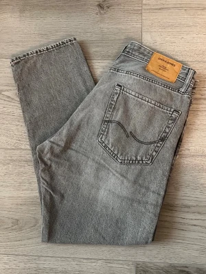 Grå jeans från Jack & Jones - Säljer ett par grå jeans från Jack & Jones med klassisk femficksdesign och raka ben. Jeansen har en snygg tvättad look och är tillverkade i denim med en skön känsla. Perfekta för dig som gillar en avslappnad stil.