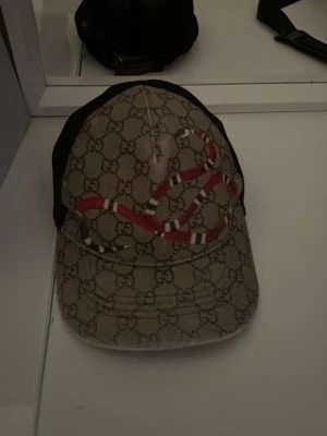 Gucci keps med orm och GG-mönster - Säljer en snygg keps från Gucci med klassiskt GG-monogram i beige och brunt samt en röd orm som detalj på framsidan. Kepsen har svart mesh på sidorna och justerbar rem bak. Perfekt för dig som vill sticka ut med en lyxig accessoar.