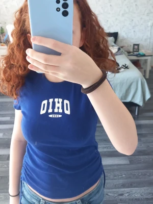 Blå OHIO t-shirt från H&M - Säljer en blå t-shirt från H&M med vit OHIO-tryck framtill. T-shirten har korta ärmar och snörning i sidan för en snygg detalj. Materialet är mjuk bomull och passformen är tight. Perfekt för dig som gillar college-vibe och sportig stil.
