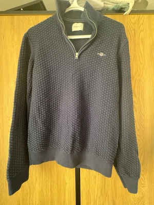 Marinblå halvzip tröja från GANT - Säljer en marinblå stickad tröja från GANT med halvzip och ribbade muddar. Tröjan har ett diskret GANT-logo broderat på bröstet och ett snyggt strukturmönster i tyget. Perfekt för dig som gillar klassisk och stilren design.