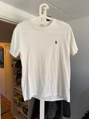 Vit t-shirt från Polo Ralph Lauren - Klassisk vit t-shirt från Polo Ralph Lauren med rund hals och korta ärmar. T-shirten har den ikoniska lilla marinblå logotypen broderad på bröstet. Tillverkad i mjuk bomull för en skön känsla och enkel stil.